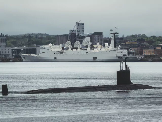 A nuclear submarine departs a harbour for the open Atlantic, while a rocket trajectory tracking ship is moored in the distance.